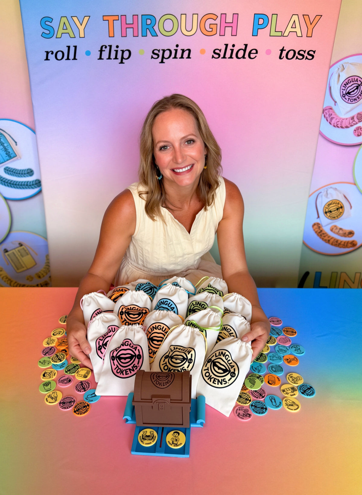 Portrait of Lisa Acosta, MA CC-SLP sitting at a table with a colorful background that says Say Through Play - roll • flip • spin • slide • toss embracing the Complete Collection of Lingua Tokens - Speech Therapy Tools and treasure chest trunk token dispenser - patent pending.